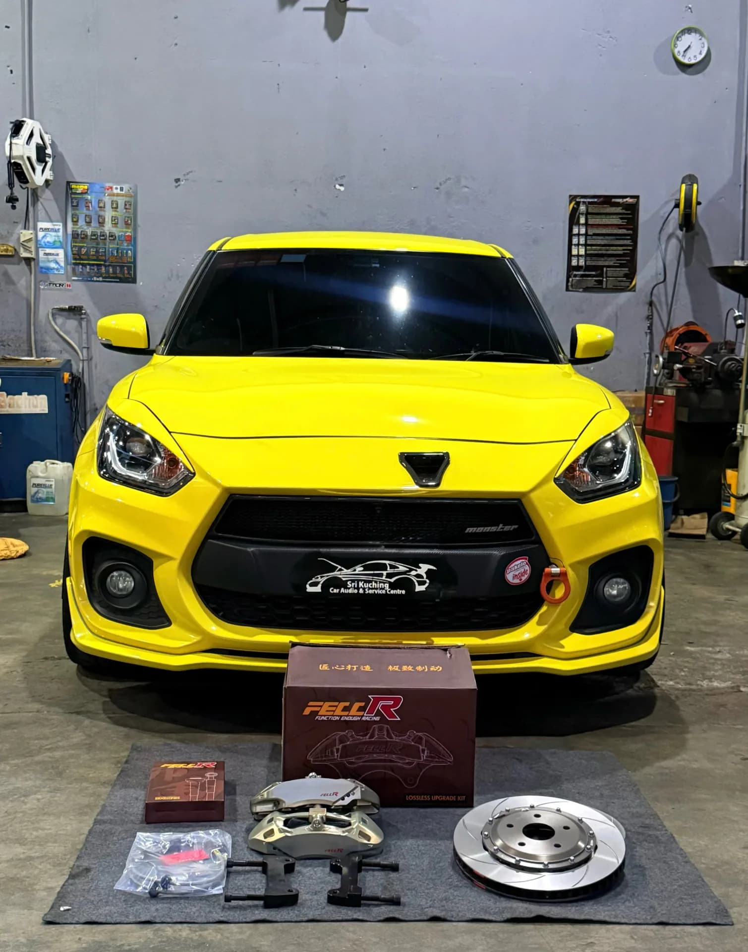 Yellow Suzuki Swift Sport ZC33S with a caliper, disc and brake kit packaging displayed in the workshop